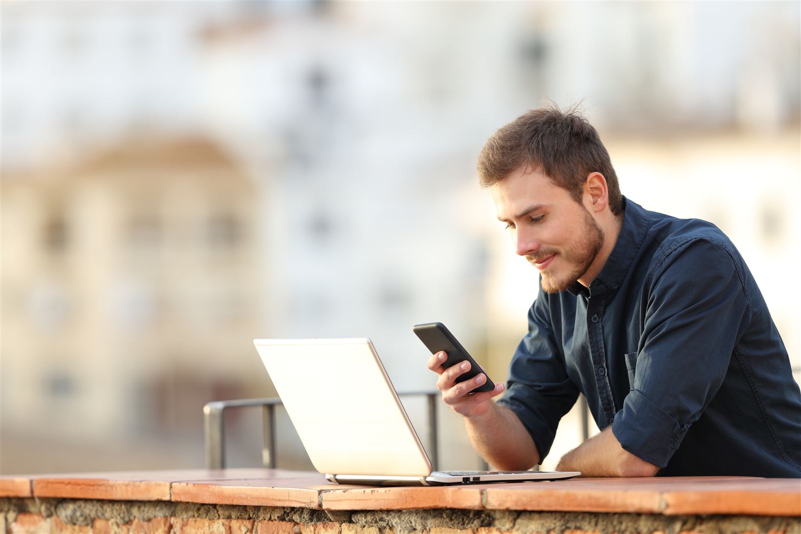 Mann arbeitet mit Laptop und Smartphone auf einem Balkon